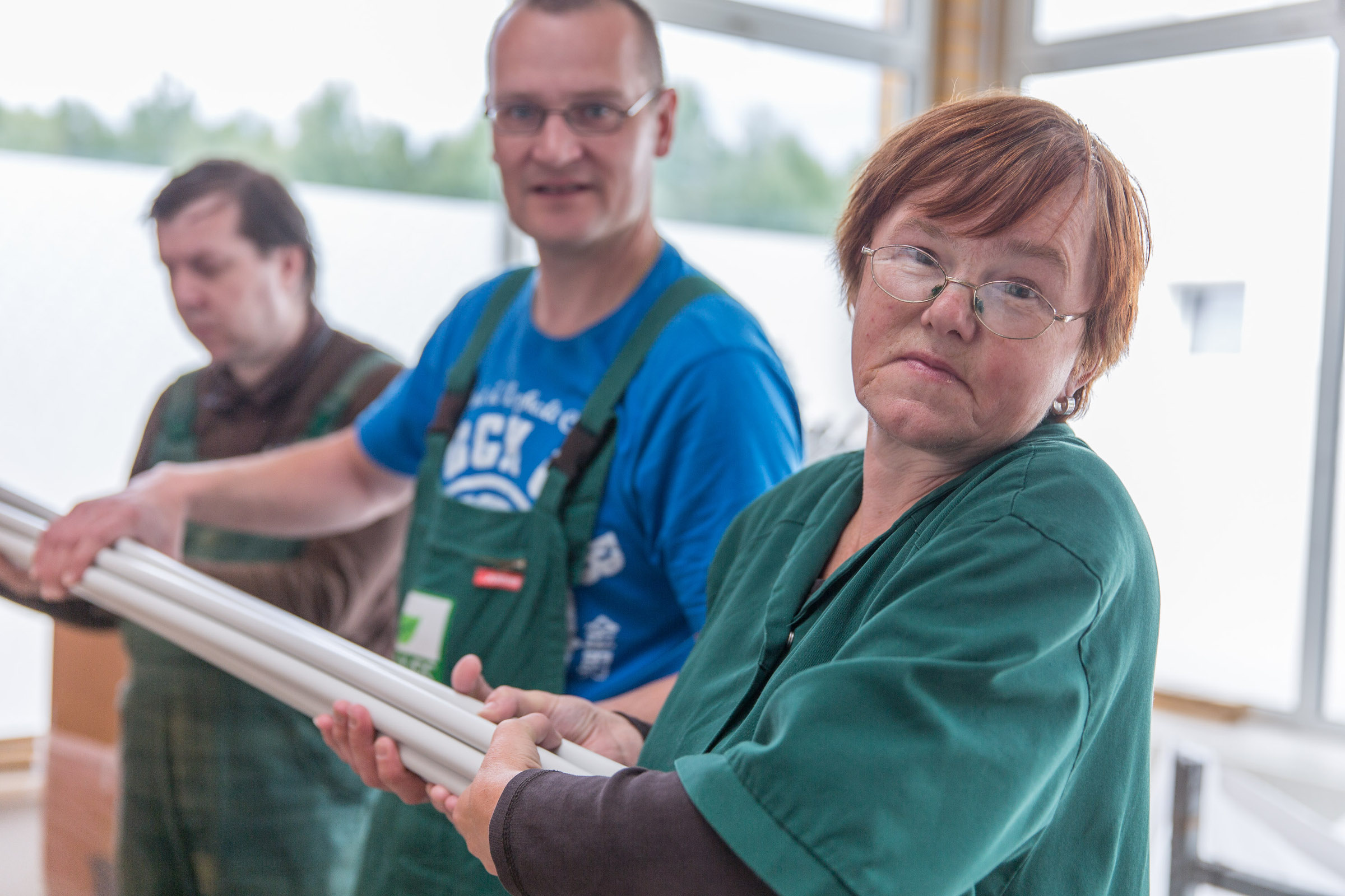 Teilhabe und Arbeit Eine Frau und zwei Männer heben mehrere Stangen gemeinsam auf. Sie tragen Arbeitskleidung und gucken in die Kamera.